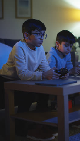 Vertikales-Video-Von-Zwei-Kleinen-Jungen,-Die-Zu-Hause-Spät-In-Der-Nacht-Mit-Einer-Computerspielkonsole-Im-Fernsehen-Spielen-Und-Controller-In-Der-Hand-Halten